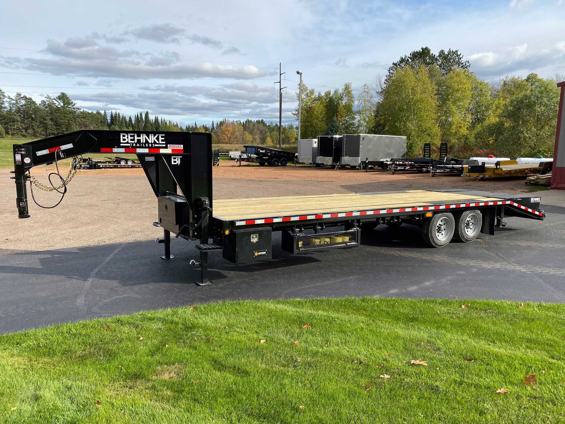 Black flatbed trailer with wooden deck, on grass in front of a lot with other trailers, under cloudy sky.