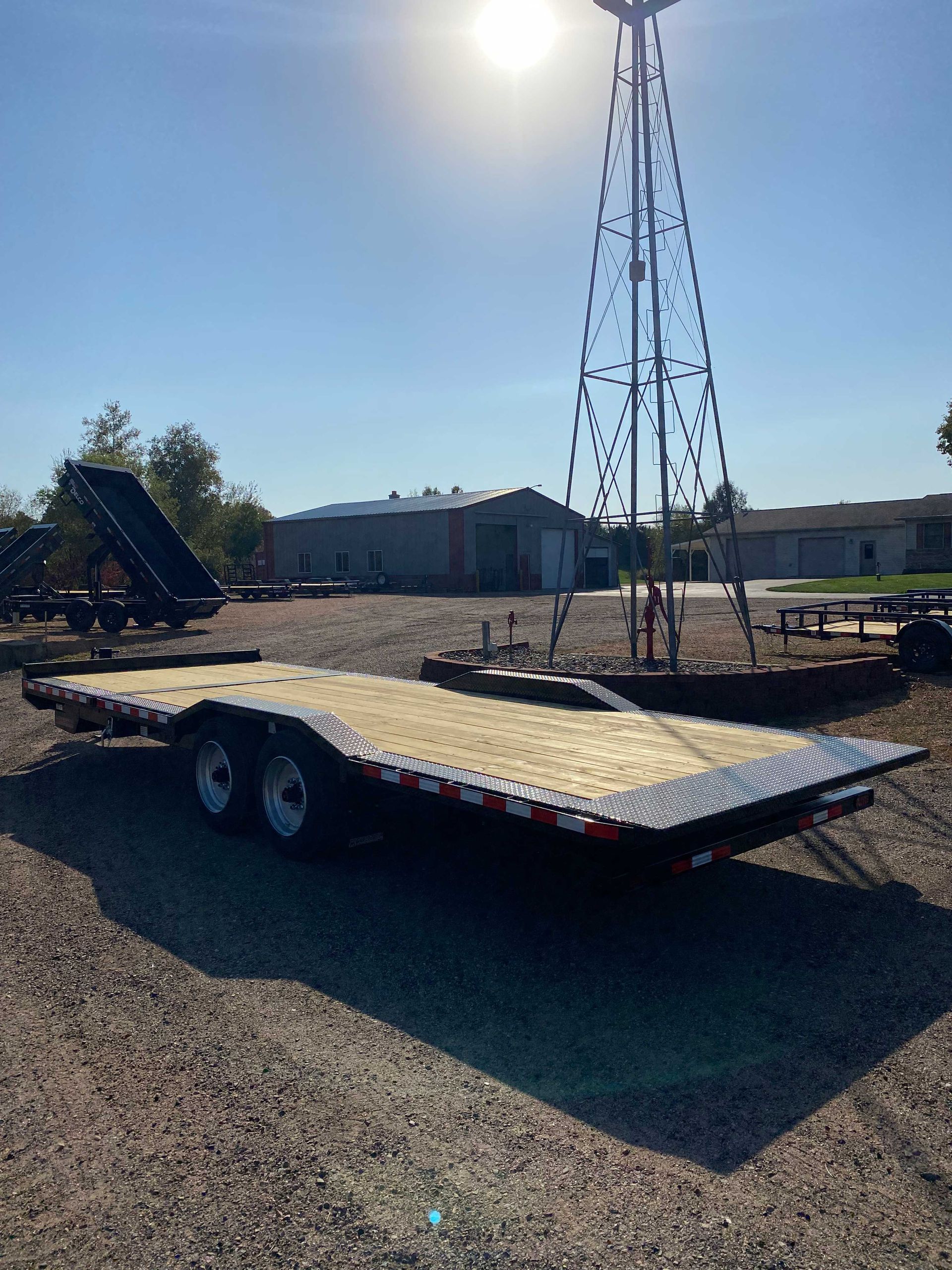 Flatbed trailer parked on gravel with a windmill and buildings in the background under a bright sun.