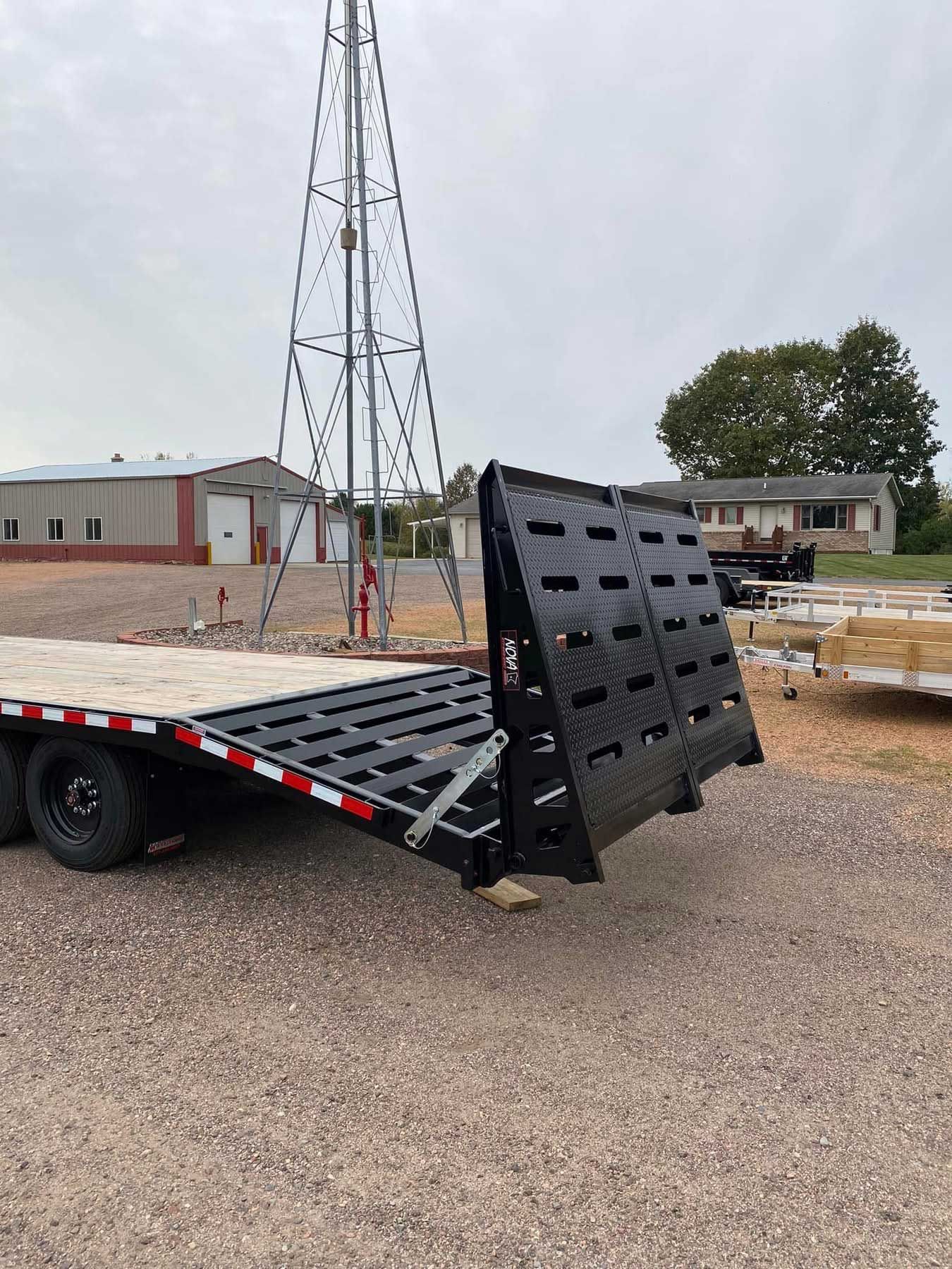 A black and red flatbed trailer with a raised ramp on a gravel lot, windmill in the background.