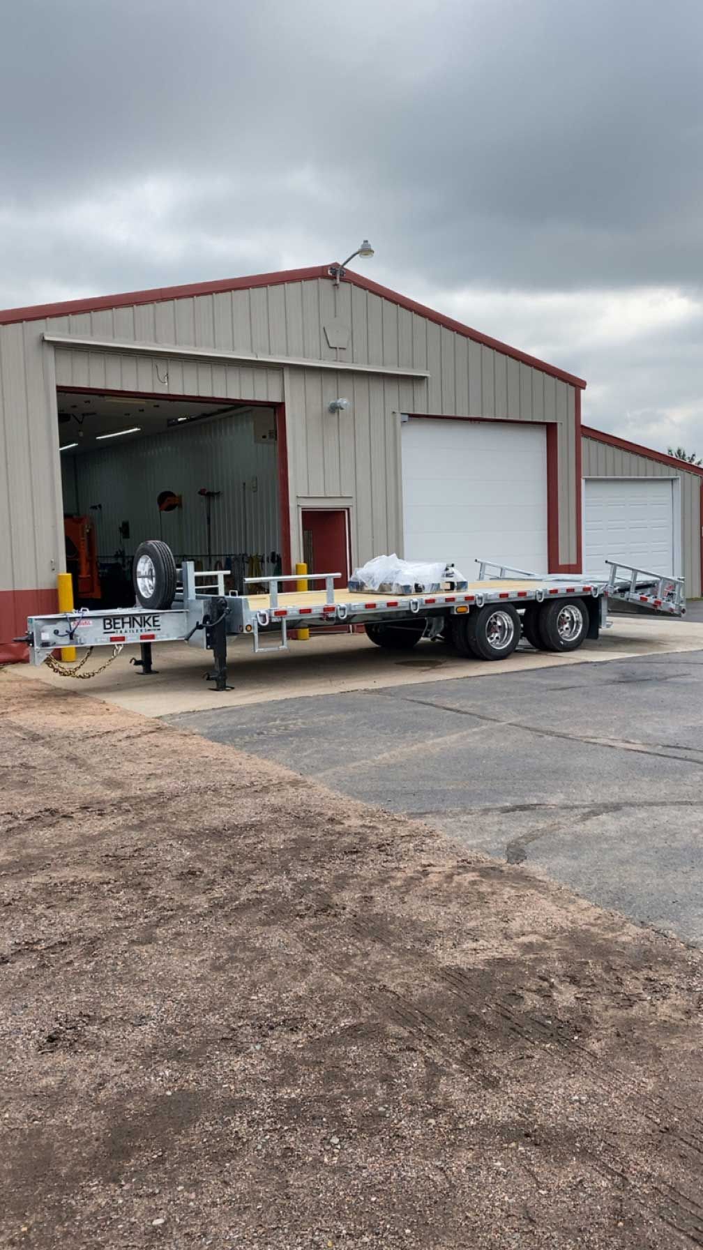 A long flatbed trailer parked outside a red and beige garage on a cloudy day.