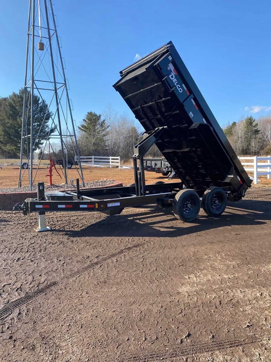 Black dump trailer with raised bed, set against a blue sky, dirt ground, and a windmill.