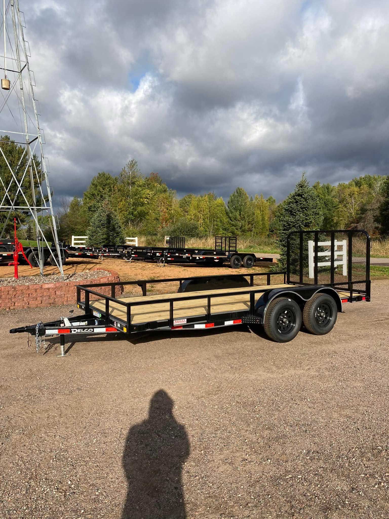 Flatbed trailer with black frame, wooden deck, and black tires parked on gravel under a cloudy sky.