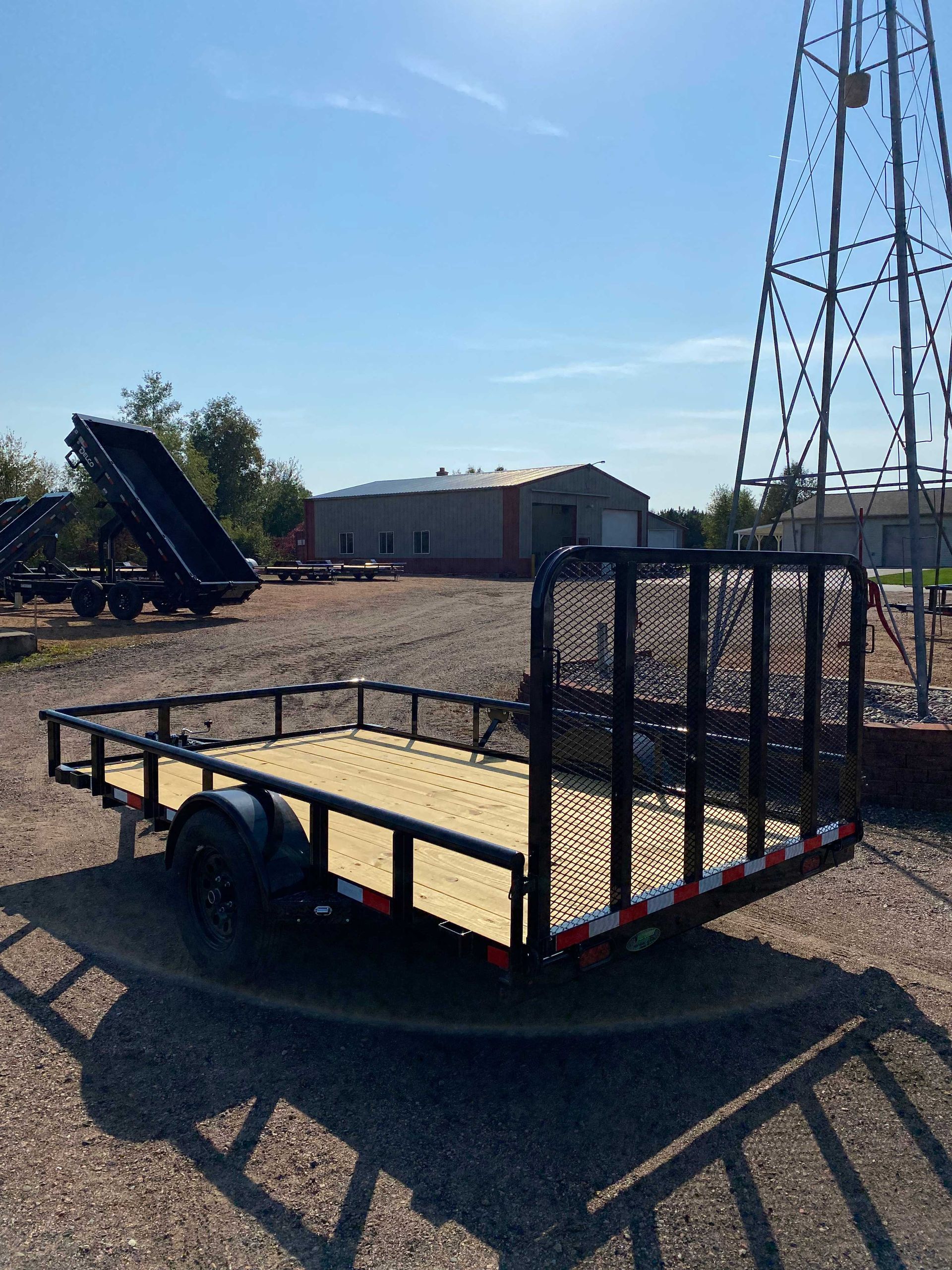 Black utility trailer with wooden deck, rear gate, parked in front of a metal tower and building, sunny outdoor setting.