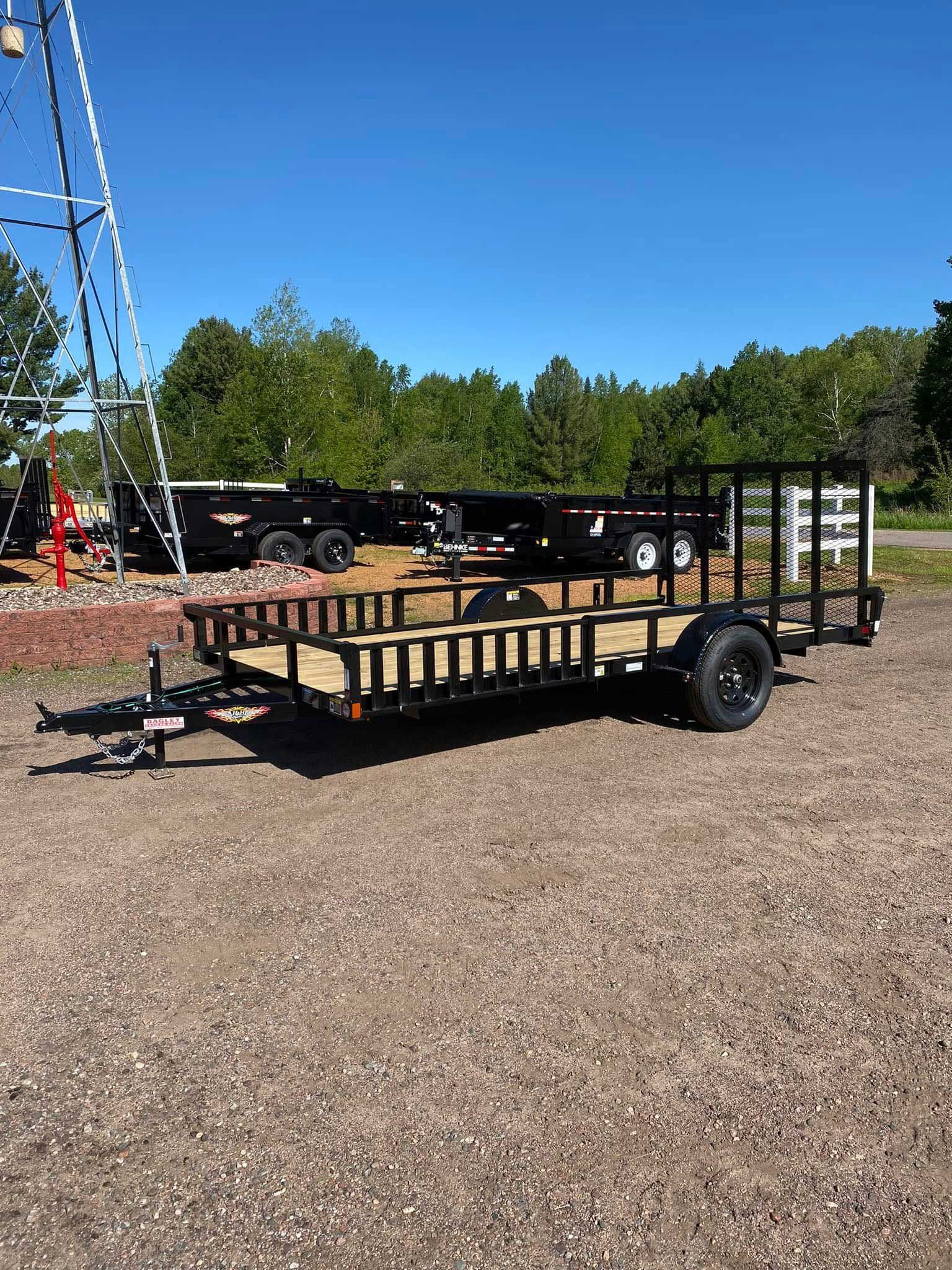 Black utility trailer with wooden bed on gravel, in front of other trailers under a blue sky.