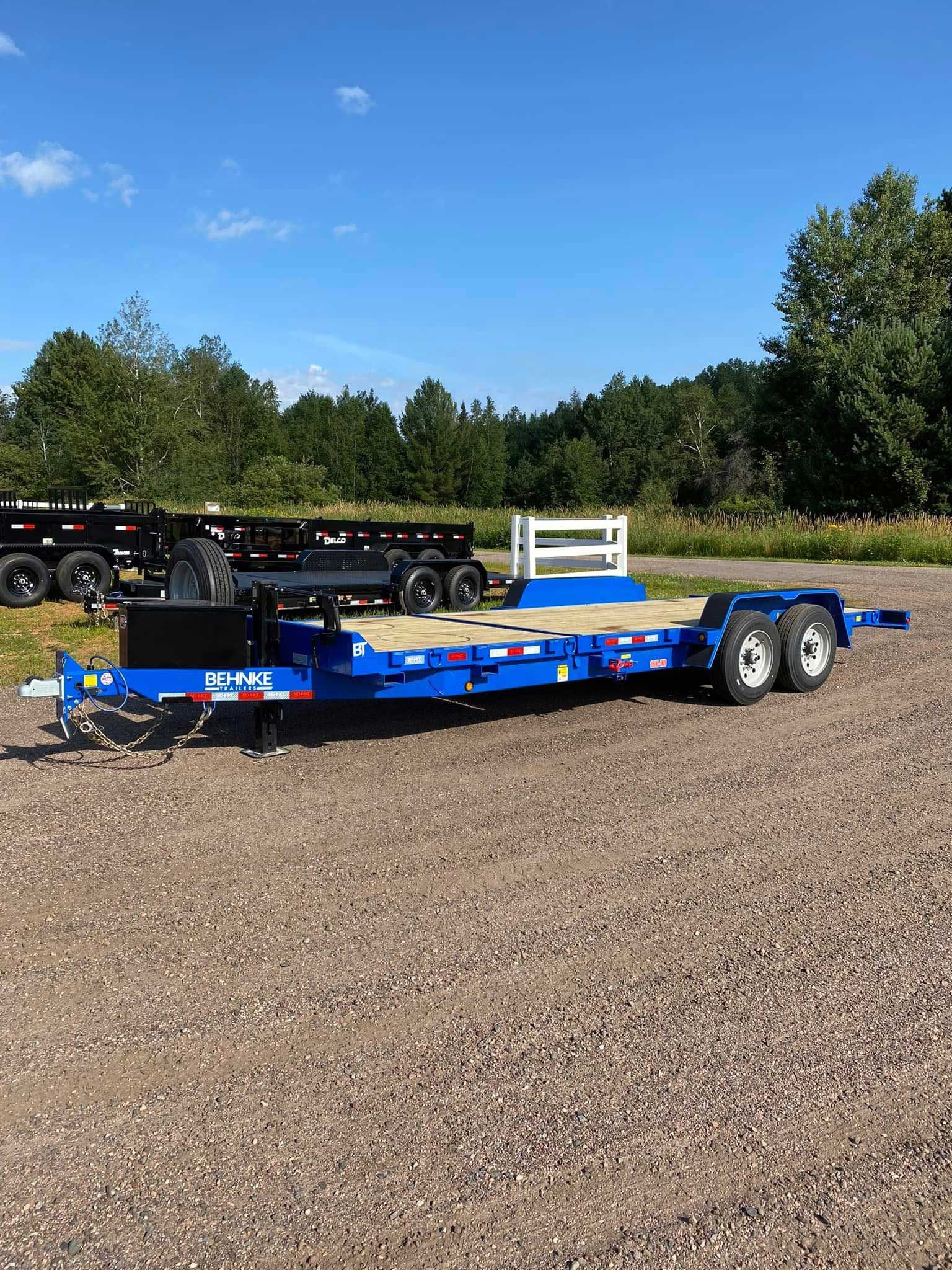 Blue flatbed trailer on gravel, against a backdrop of trees and a blue sky.