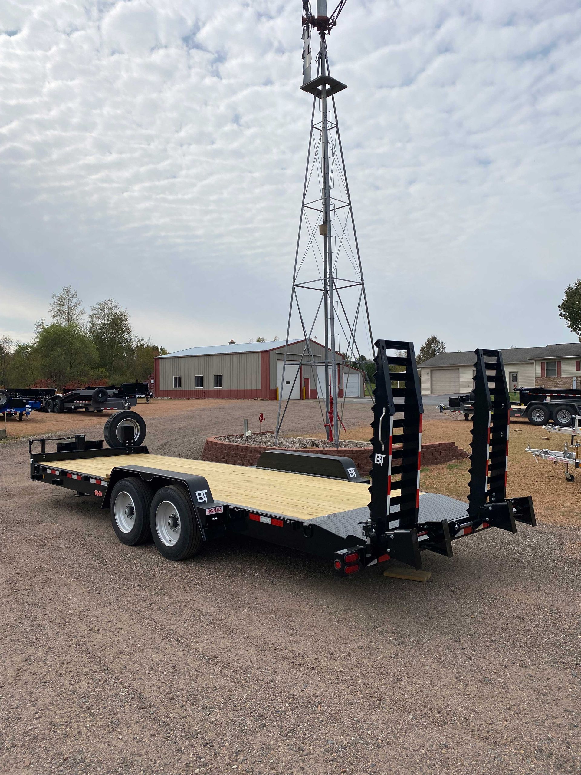 Black trailer with wooden deck, ramps, and dual axles. Standing in a gravel lot with a windmill in the background.