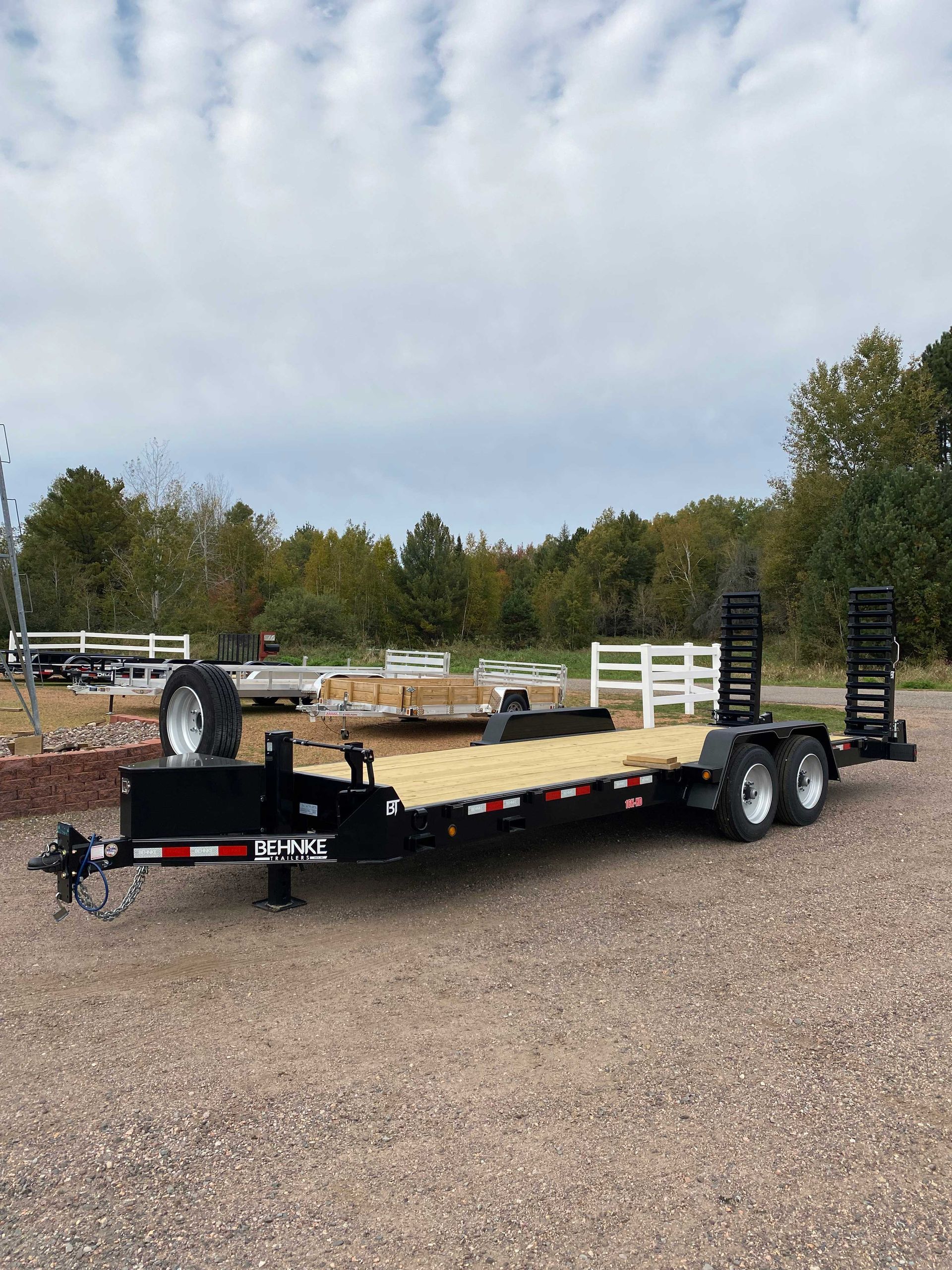 Black flatbed trailer with wooden deck, dual axles, and ramps on gravel in outdoor setting.