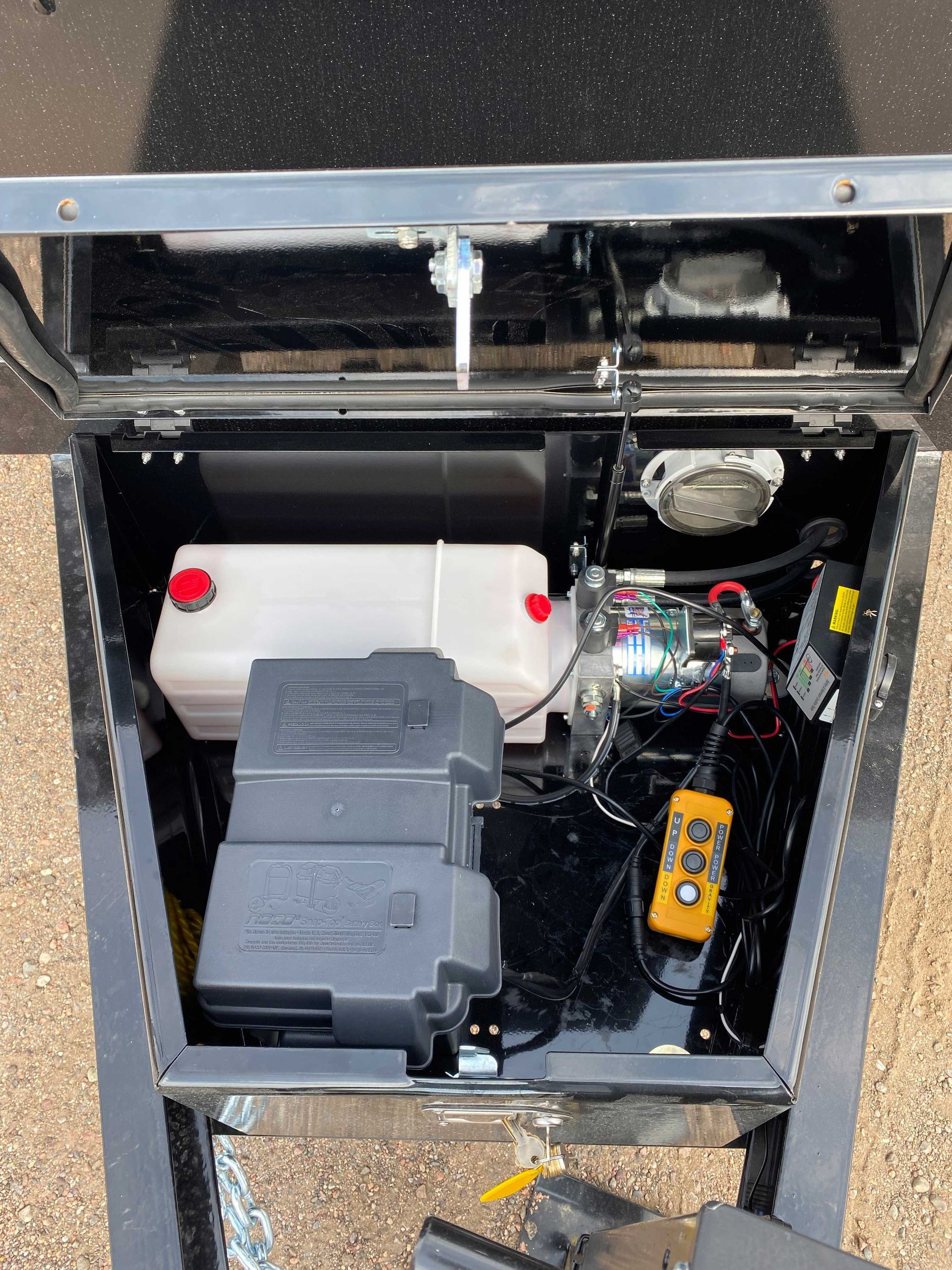 Open compartment on a trailer with a hydraulic pump, battery, and fluid reservoir. Black and silver components visible.