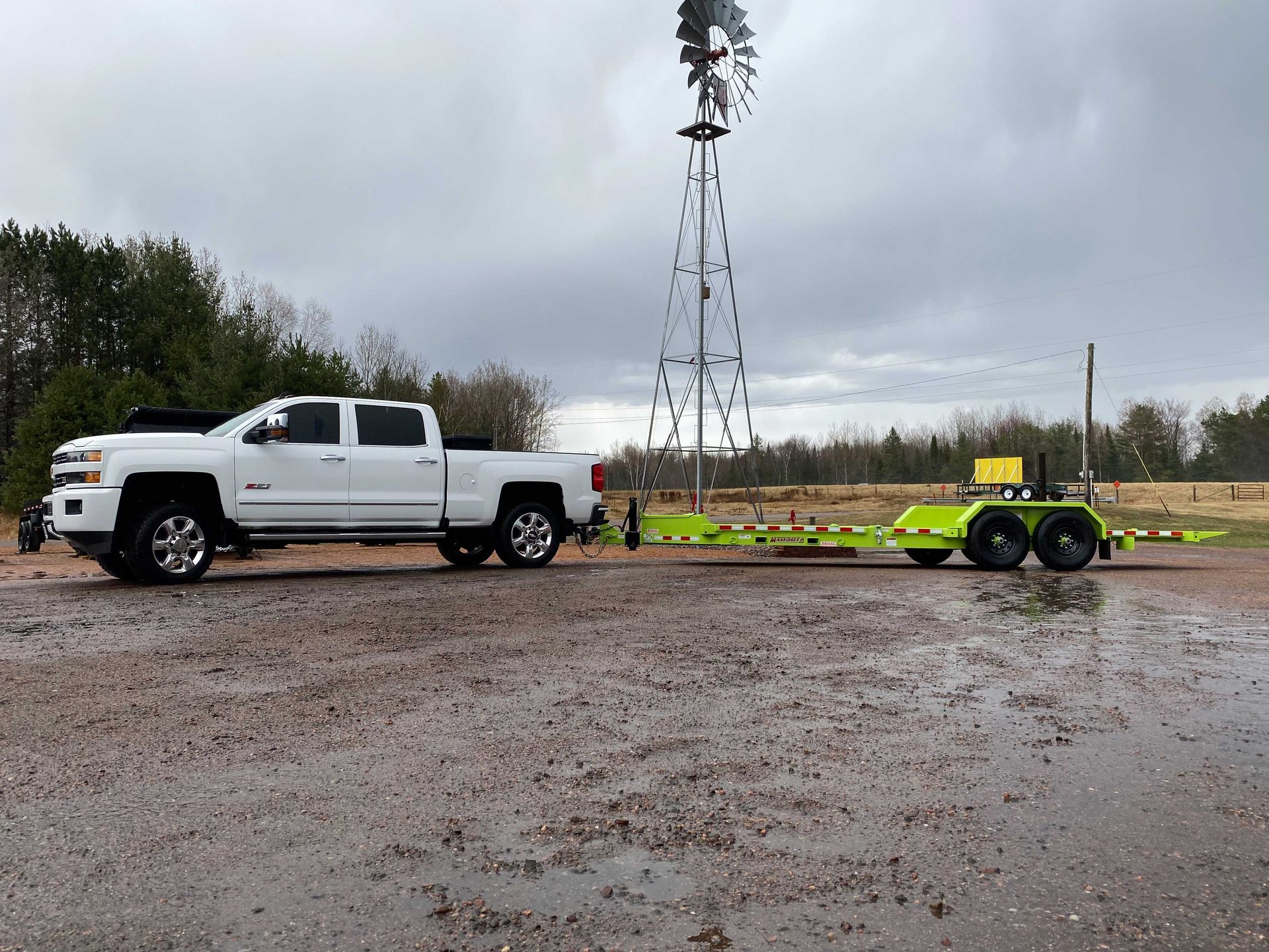 White pickup truck towing a green trailer near a windmill in a rural setting.