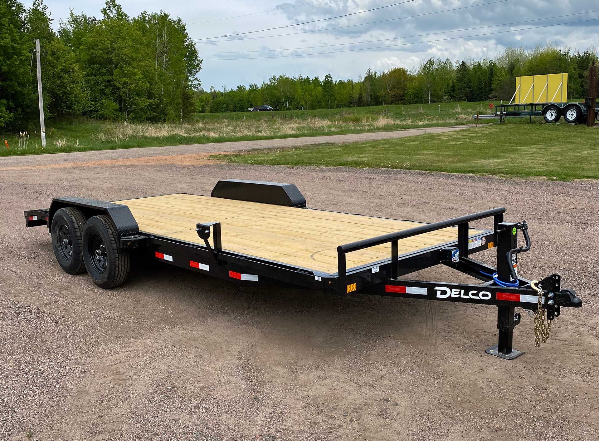 Black and red Delco flatbed trailer parked on gravel, with a wooden deck and a black frame.