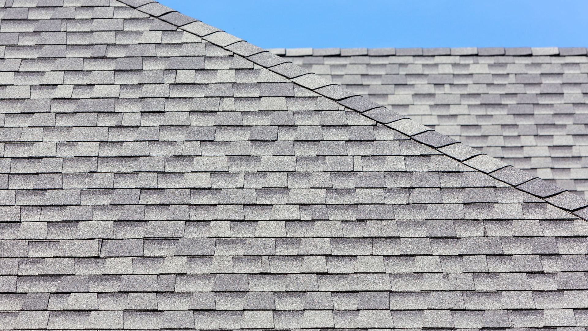 A close up of a gray shingle roof with a blue sky in the background.