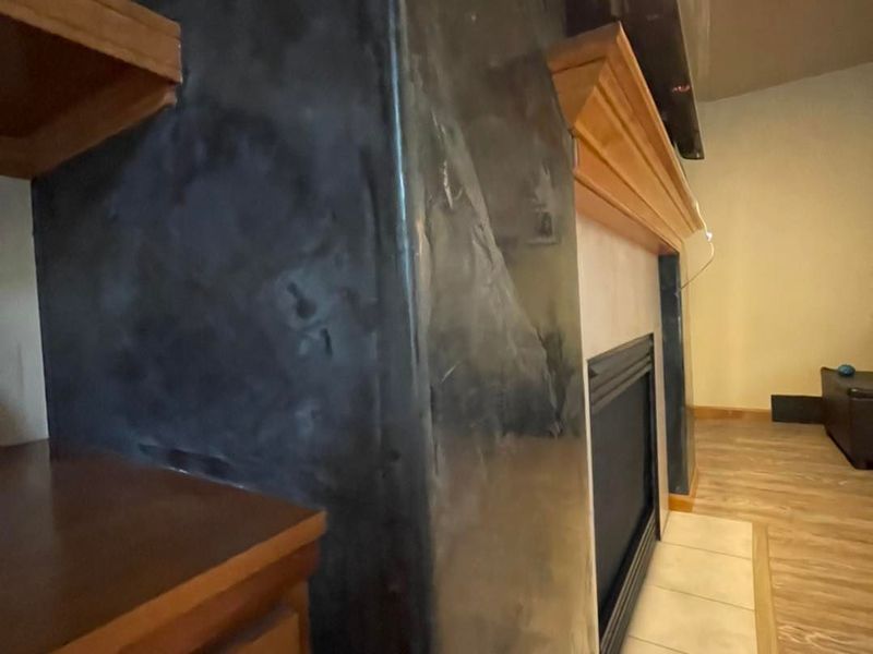 Fireplace with dark textured surround and wooden mantel, near a wooden shelf.