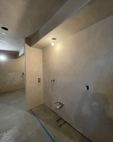 Interior of a room under construction, with beige walls, concrete floor, electrical outlets, and a light fixture.