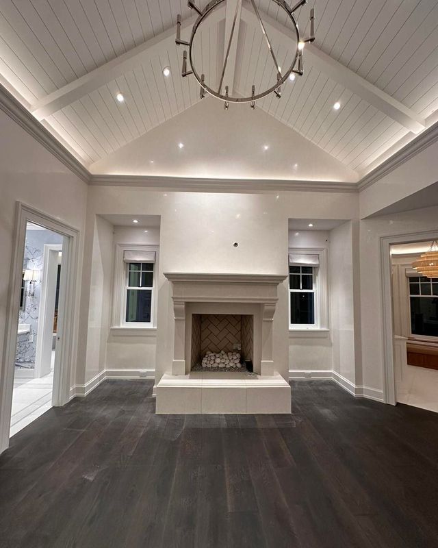 Spacious room with fireplace, white walls, dark wood floor, windows, and a vaulted, paneled ceiling.