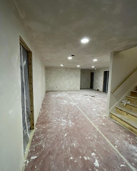 Unfinished basement with drywall, recessed lights, and a staircase. Floor covered in protective sheeting.