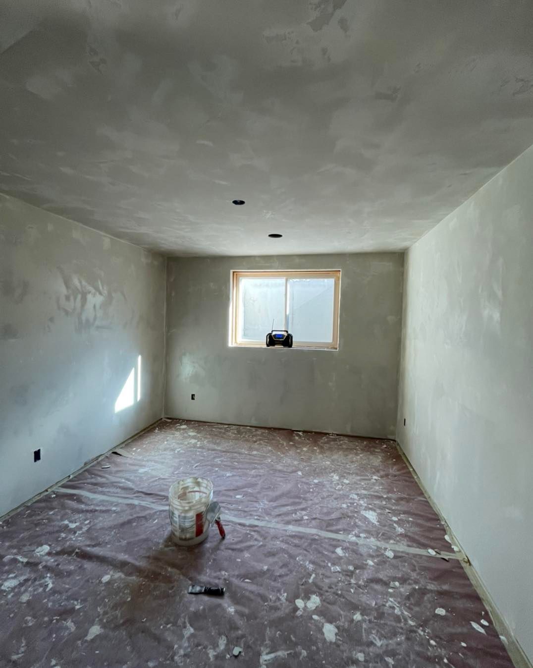 Room under construction; walls and ceiling plastered, window centered, floor covered in protective sheeting, bucket with tools.