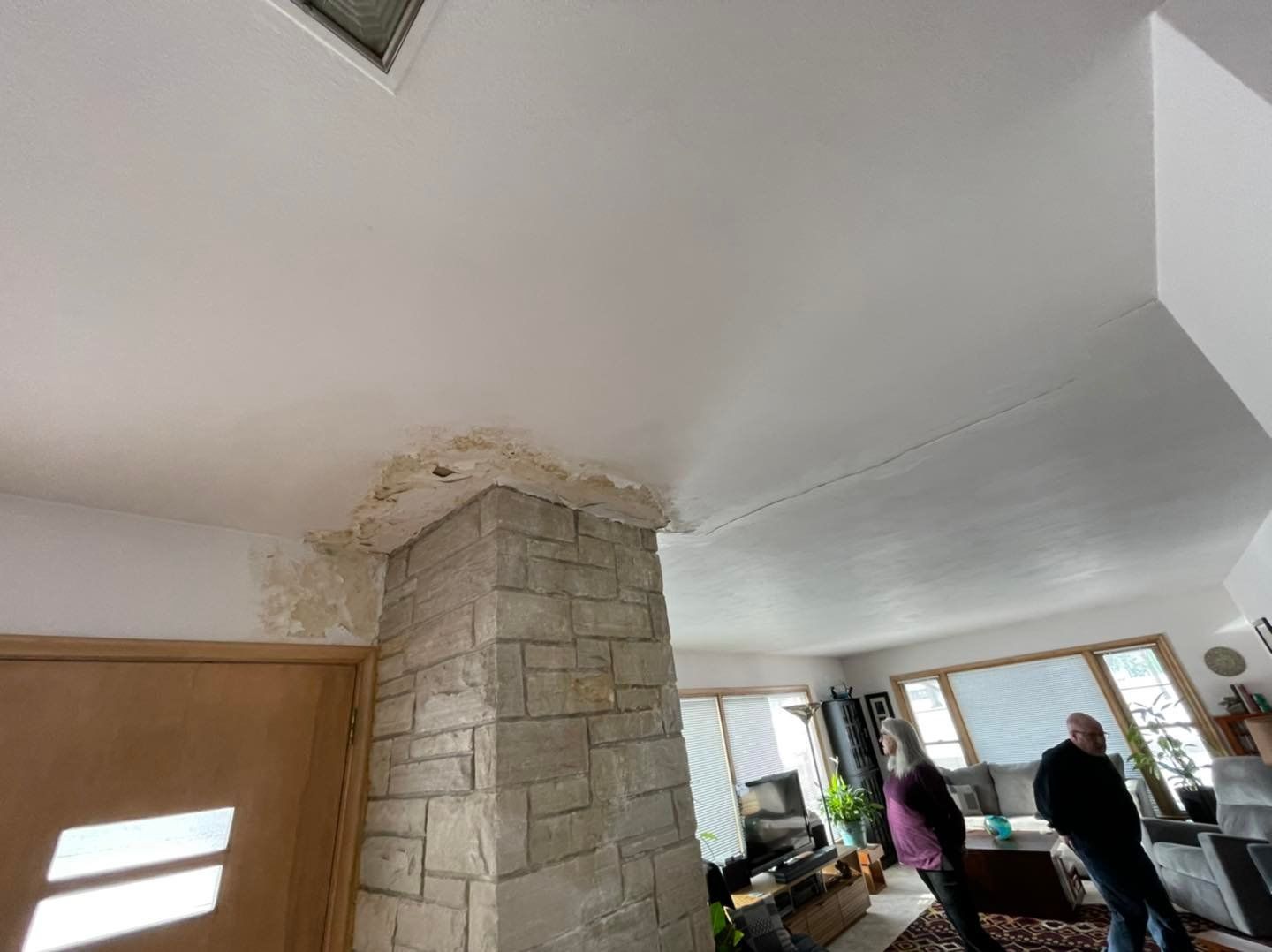 Ceiling with water damage near a stone-covered column and a doorway. Two people are in a room with a window.