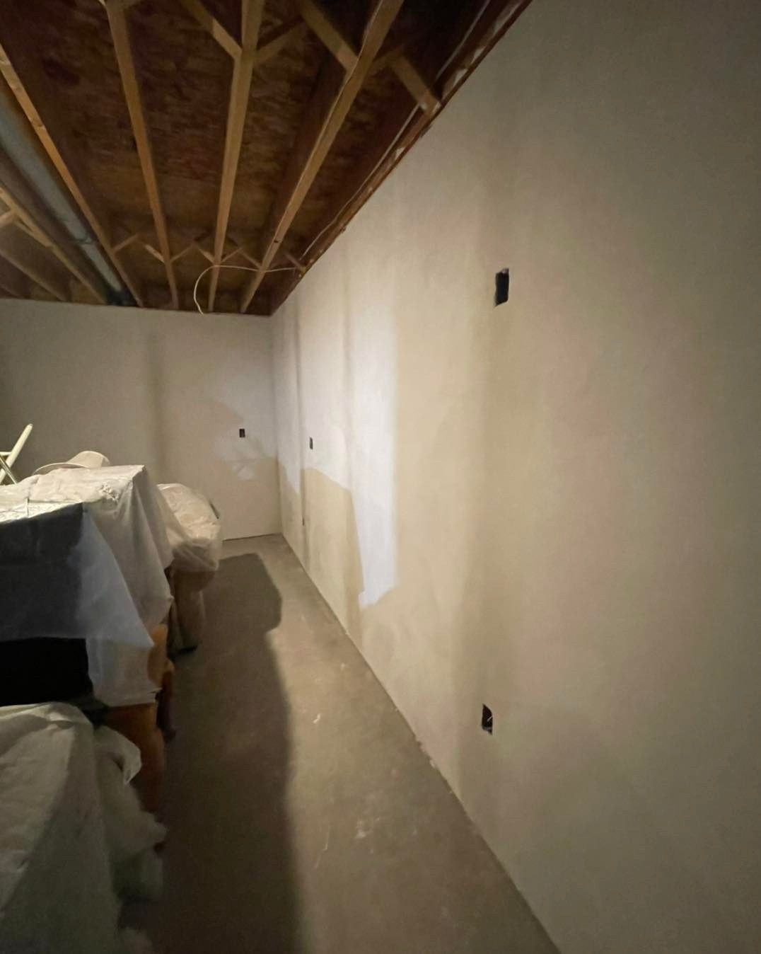 Basement room with white walls, exposed wooden ceiling, and concrete floor. Tables are covered with protective sheeting.