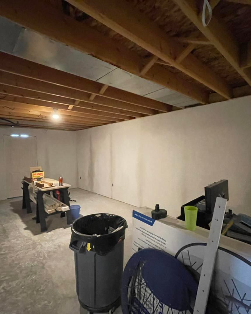 Basement with unfinished walls, exposed ceiling beams, concrete floor, worktable, trash can, and miscellaneous items.