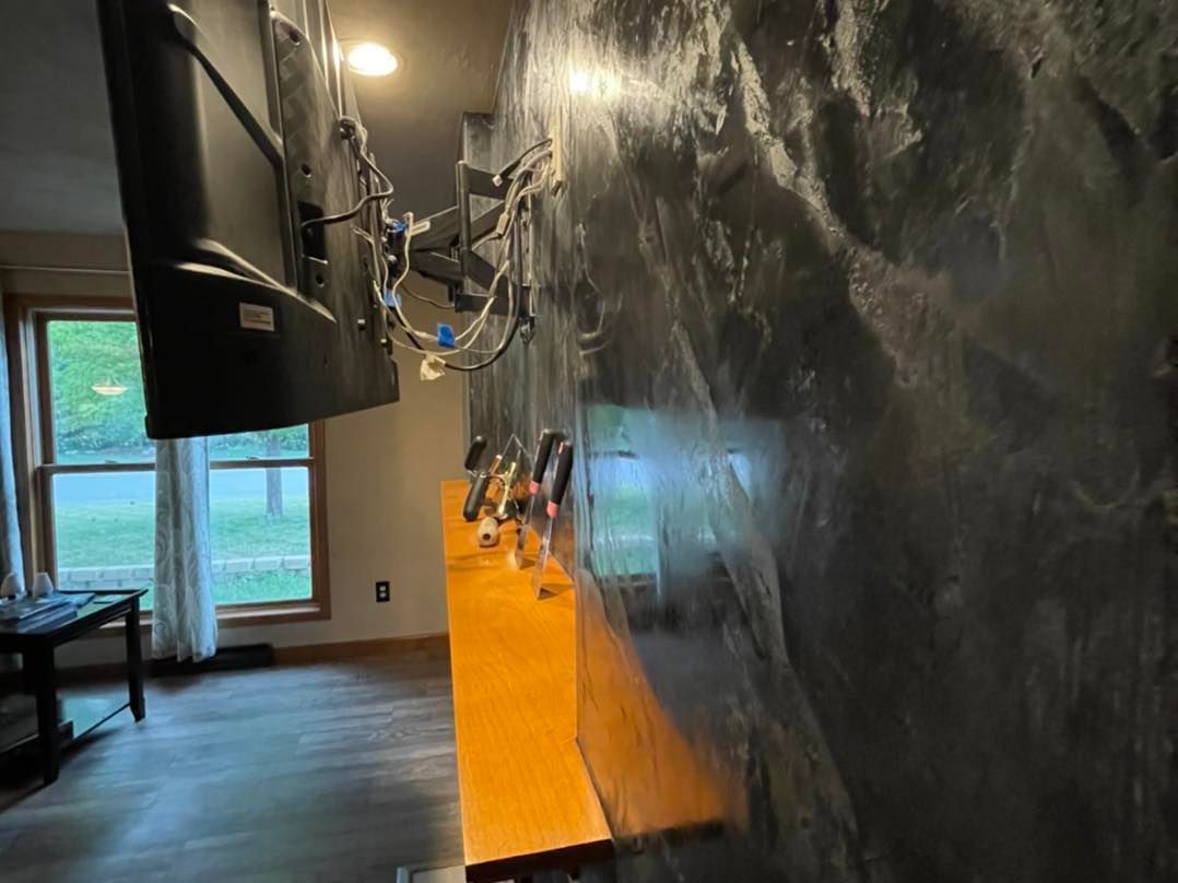 A living room with a stone accent wall, TV, and wooden cabinet. Window with view of trees is visible.