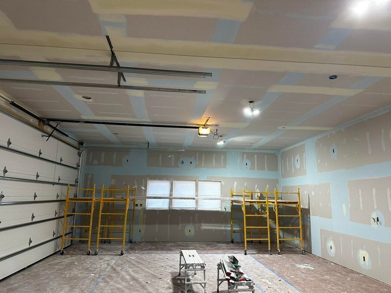 Garage interior under construction; drywall installation, scaffolding, garage door visible.