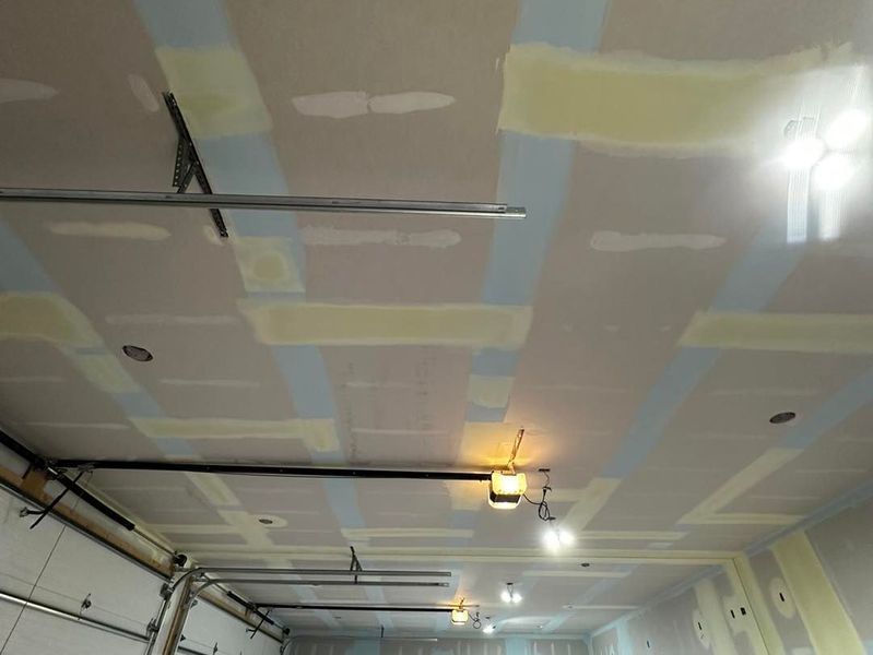 Garage ceiling with drywall, seams taped and mudded; visible overhead door tracks, lights, and wiring.