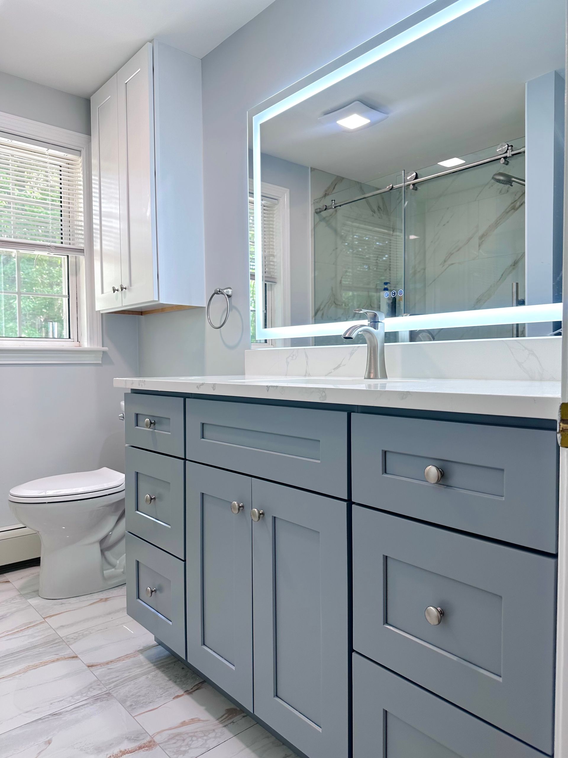 Bright bathroom with gray vanity, illuminated mirror, white countertop, and a toilet by a window.