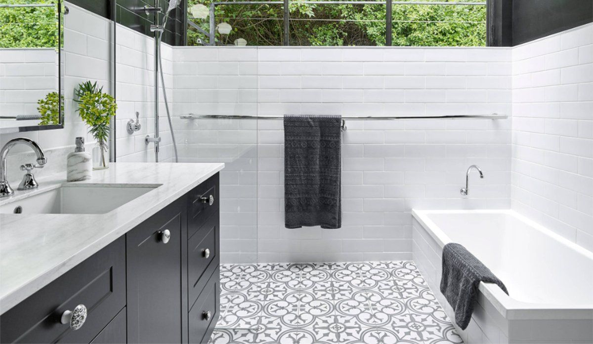 Modern bathroom with dark vanity, white tub and tiles, patterned floor, and a window with greenery outdoors
