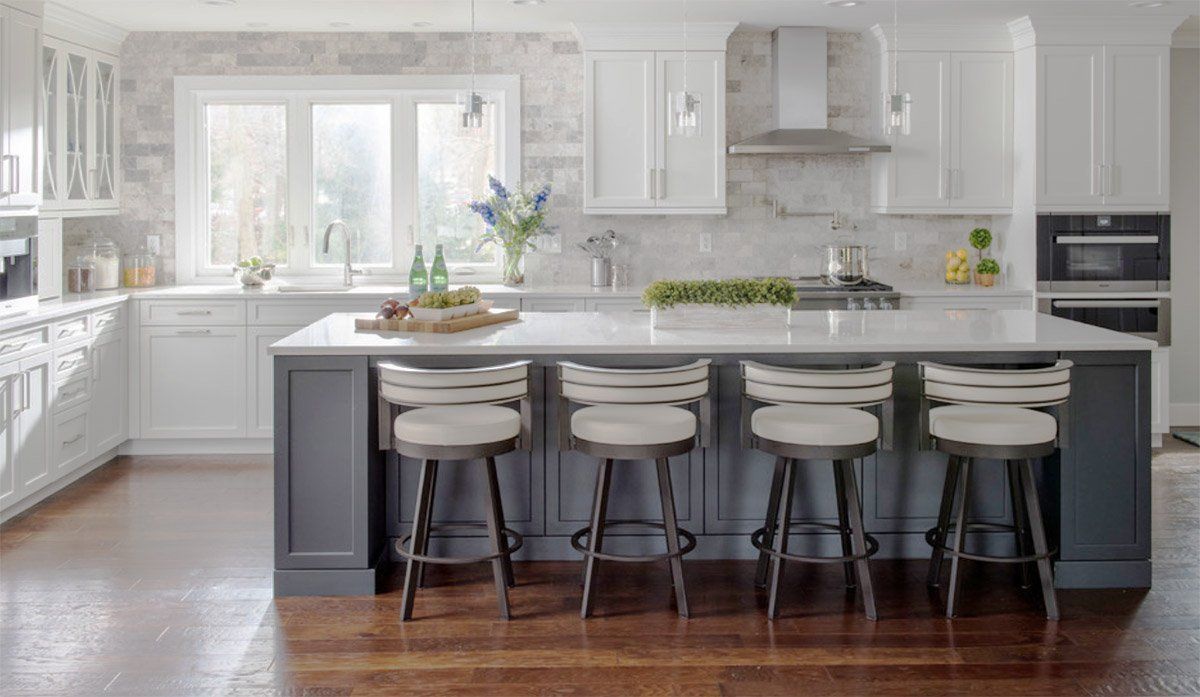 Bright white kitchen with a large island, gray stools, hardwood floors, and flowers by the window.