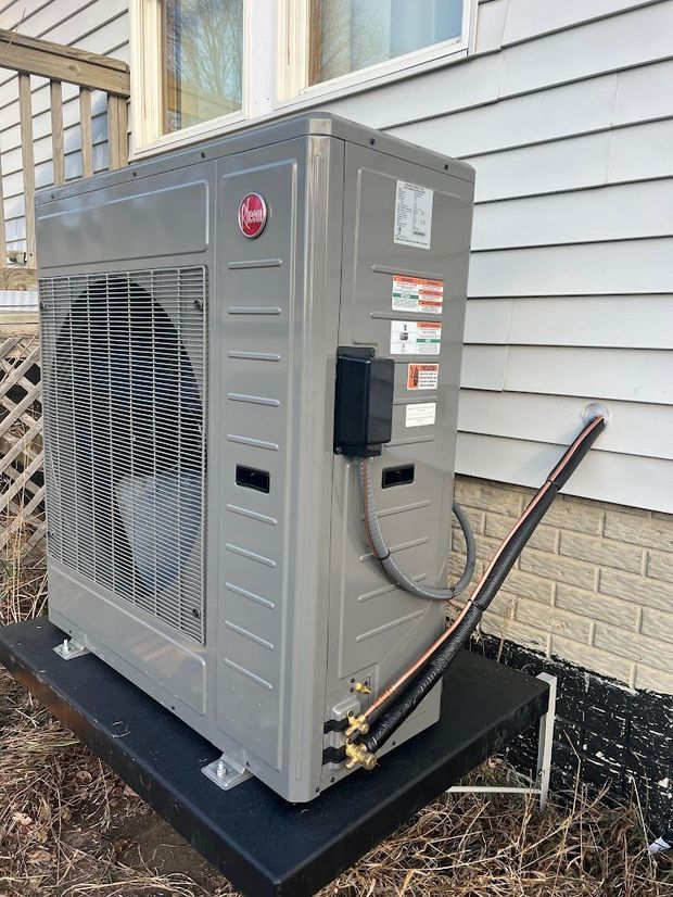 Rheem heat pump unit on black platform next to a house. Piping visible.