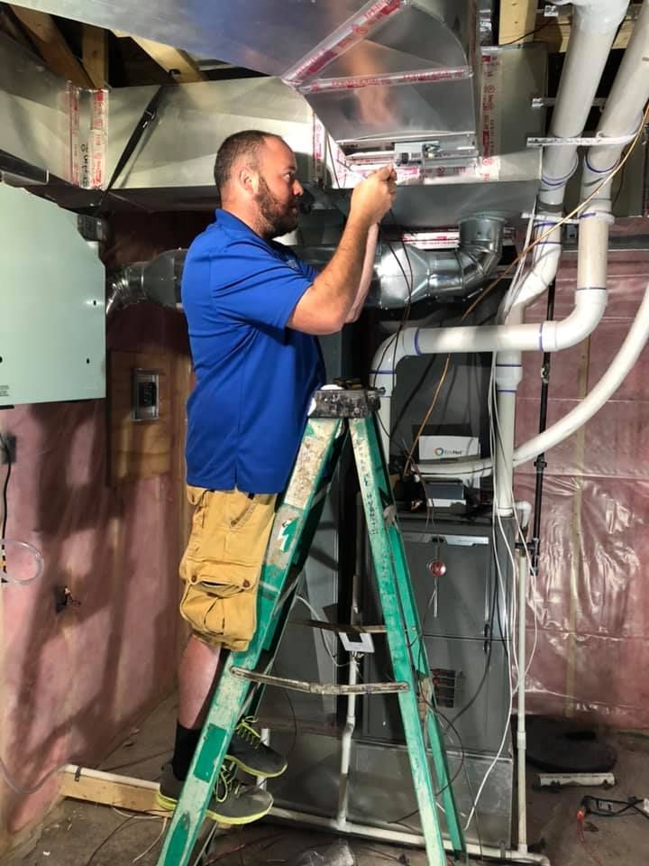 HVAC technician in blue shirt and khaki shorts on a ladder, working on ductwork near a furnace.