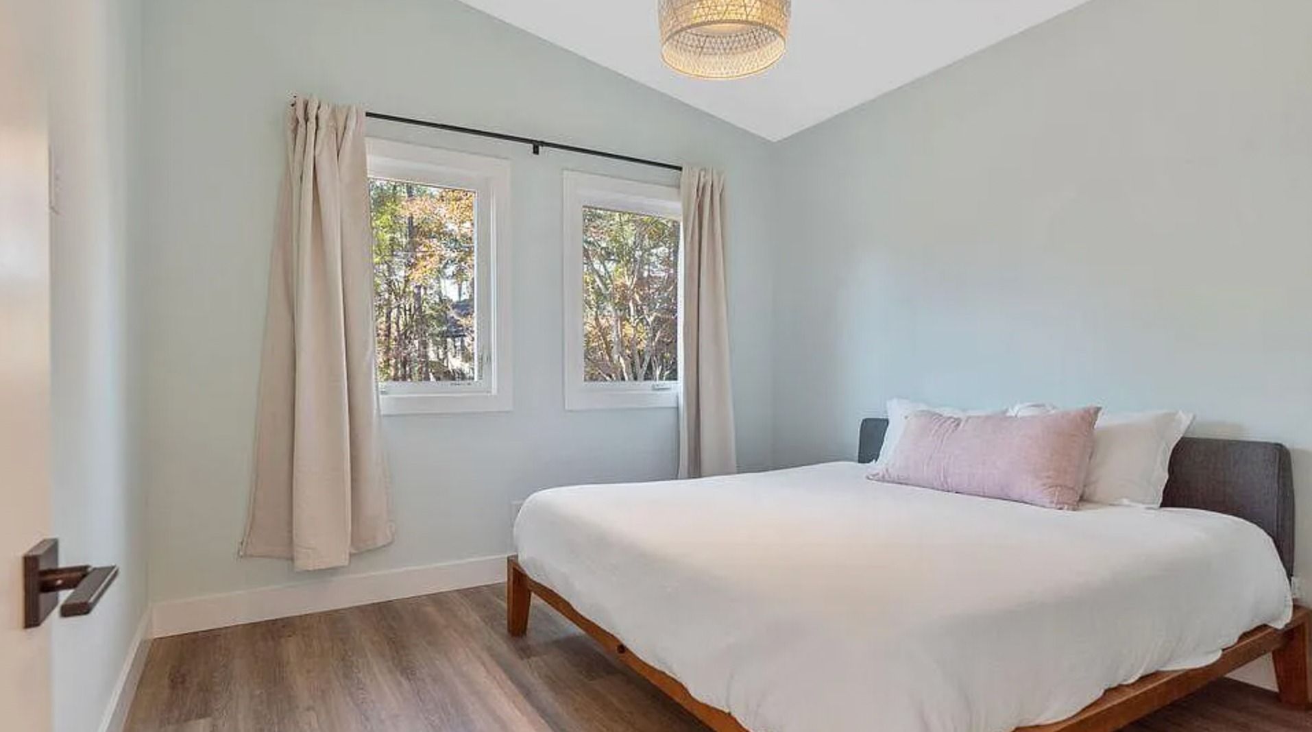 Bright bedroom with a white bed, light curtains, wood floor, and a window looking onto trees.