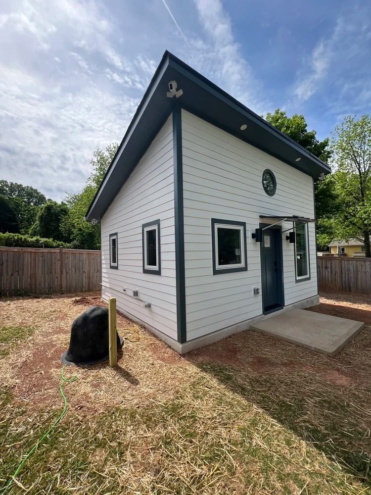 Small white house with black trim and steep roof beside a wooden fence and gravel yard