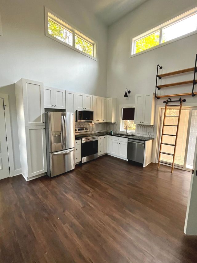 Bright kitchen with white cabinets, stainless appliances, dark wood floors, and high windows in an empty room