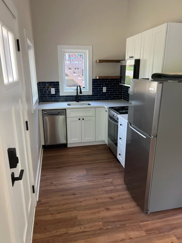 Small modern kitchen with white cabinets, black backsplash, stainless fridge, and wood flooring