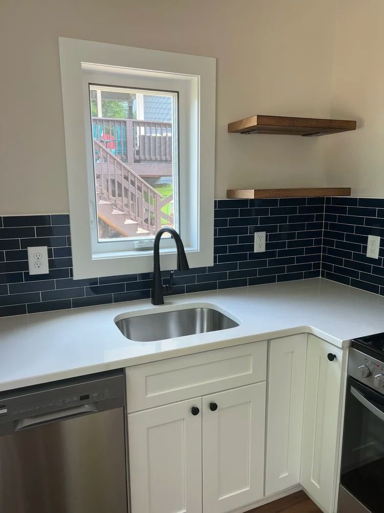 Modern kitchen sink with white cabinets, black faucet, blue tile backsplash, and window view of a deck.