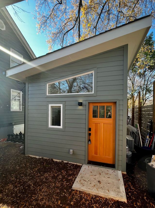 Gray house exterior with orange front door and small windows, framed by trees in daylight