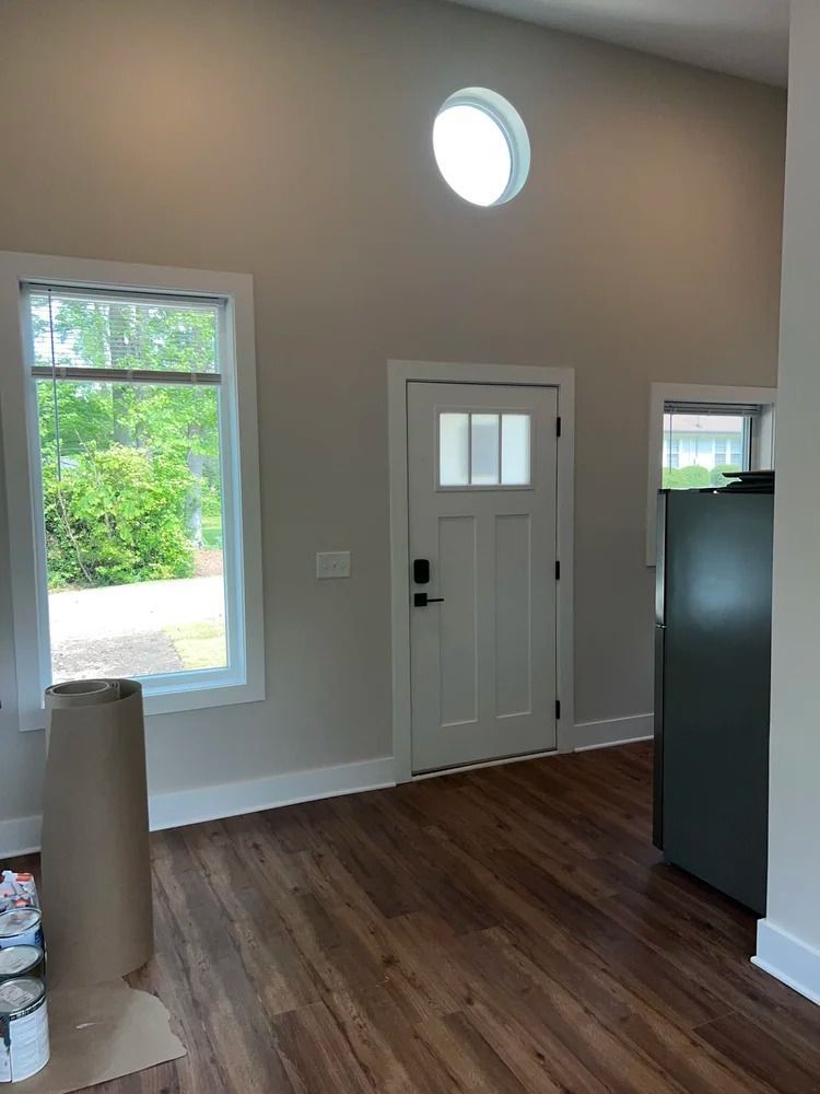 Bright empty room with wood floors, white door, tall window, and a round skylight near the ceiling