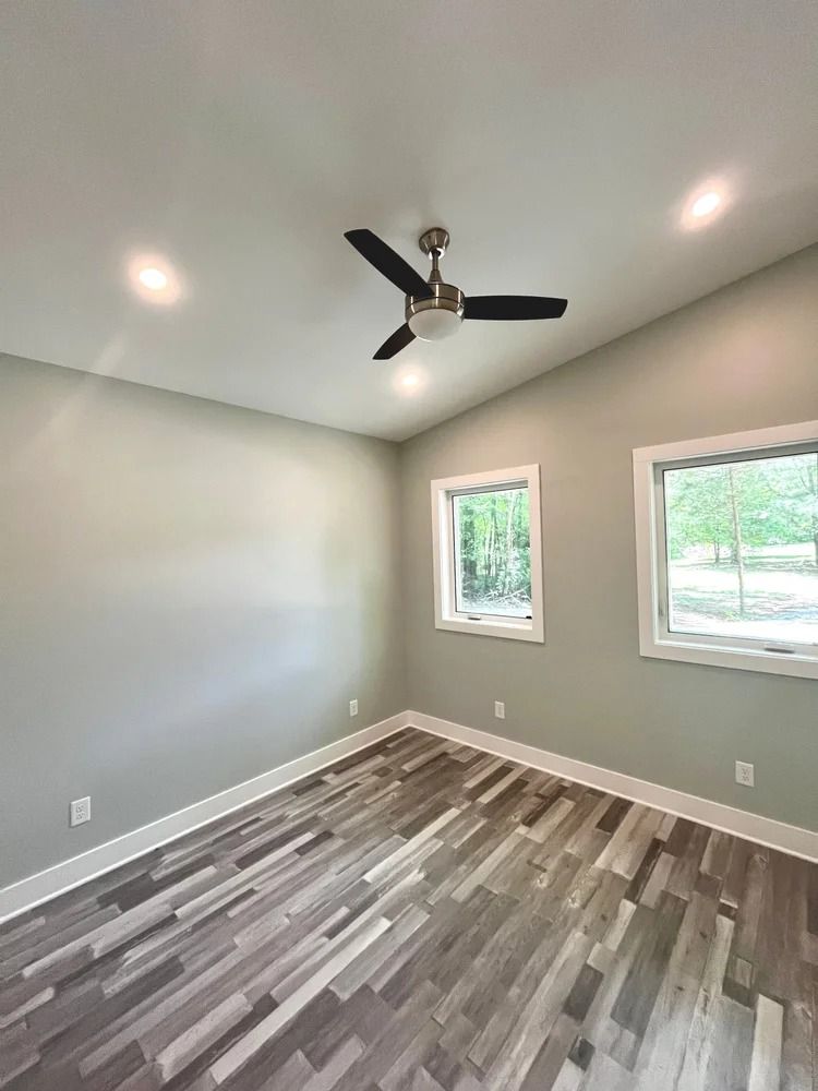 Empty bedroom with gray walls, wood-look flooring, two windows, and a ceiling fan with recessed lights