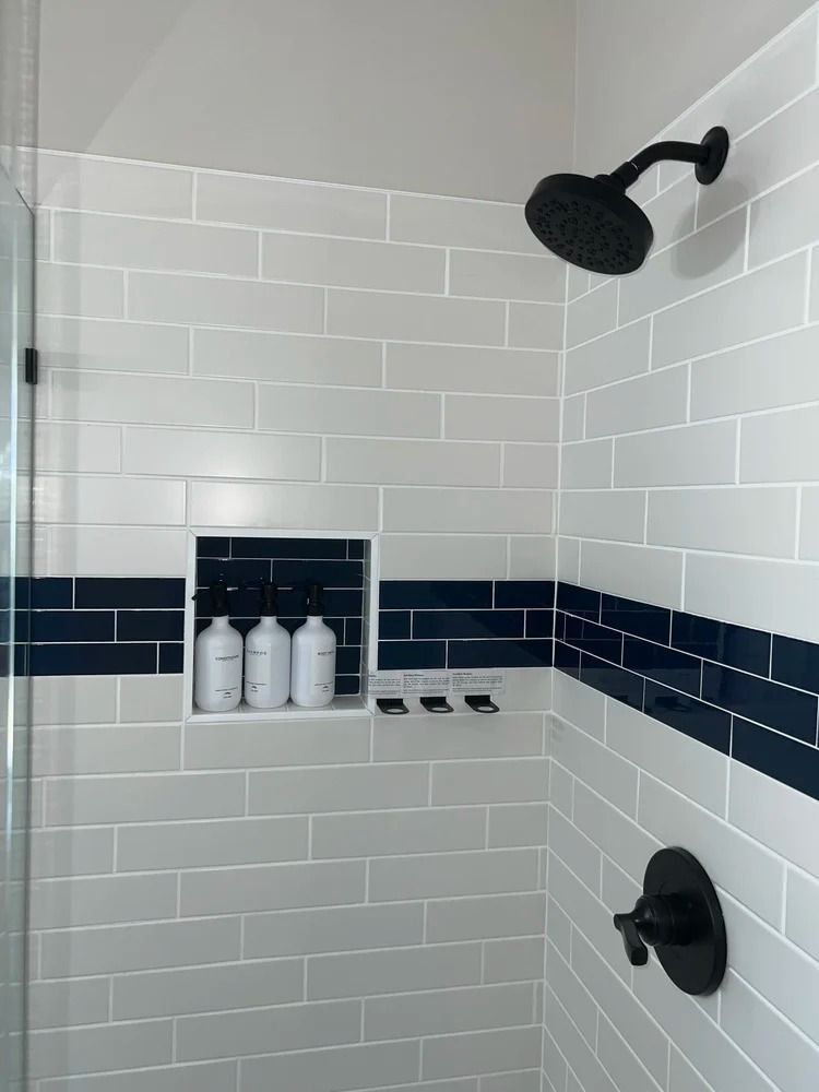 White tiled shower with black fixtures, navy accent band, and recessed shelf holding bottles.