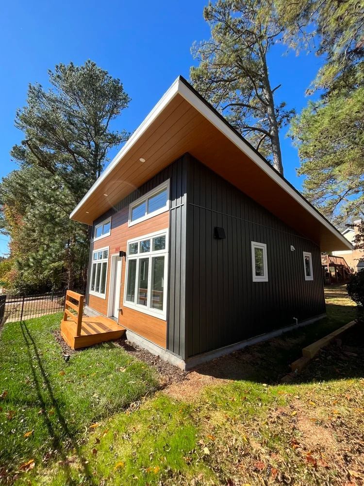Modern black cabin with wood trim and large windows in a sunny wooded yard