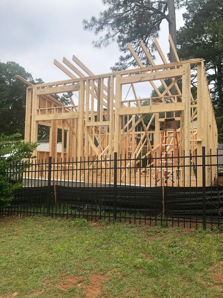 Wooden house frame under construction behind a black metal fence, with tall beams and cloudy sky.