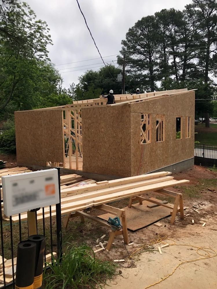 Construction site with a small wood-framed building under assembly, surrounded by lumber and tools outdoors.