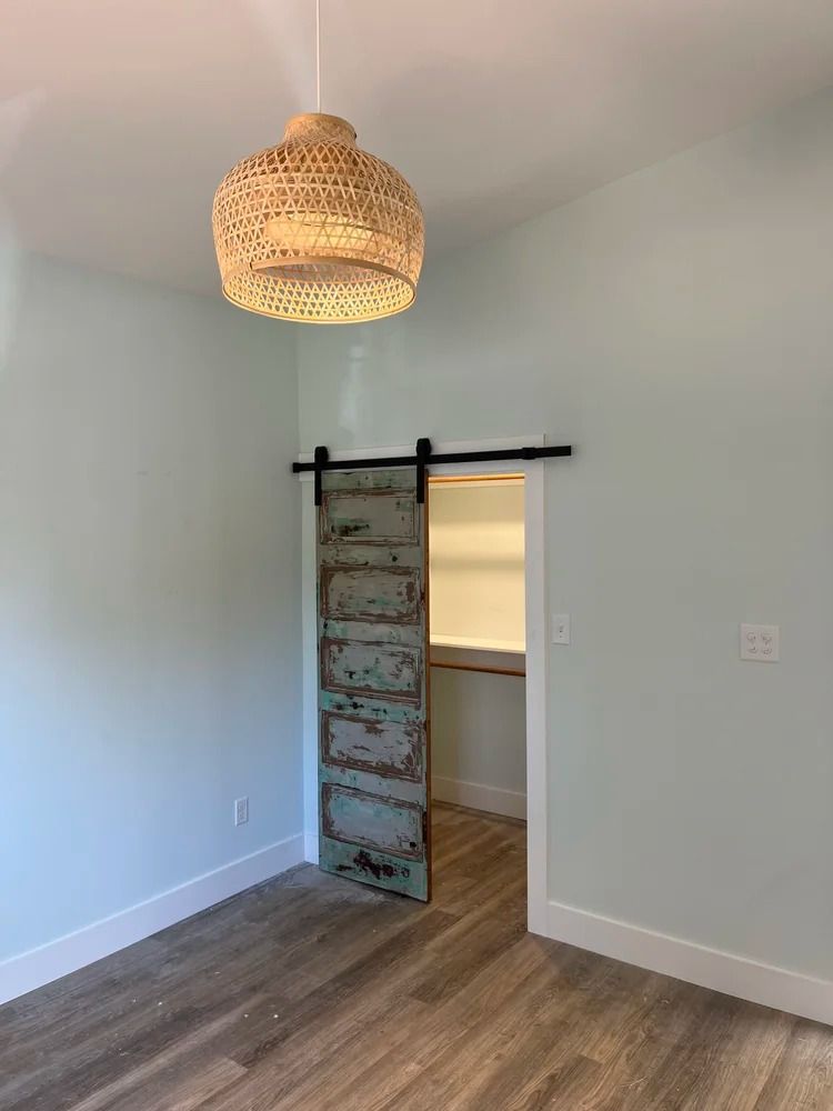 Empty room with wood floor, pale walls, woven pendant light, and a sliding barn door opening to a doorway.