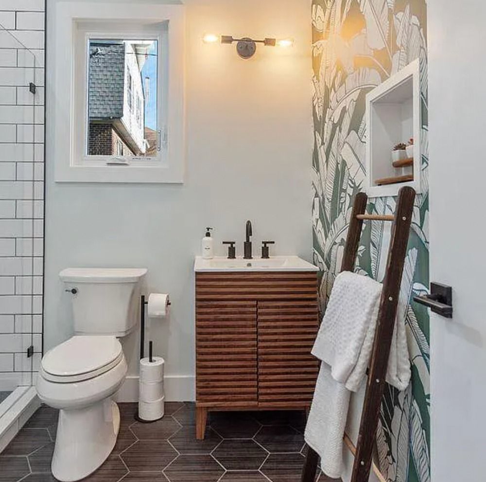 Modern bathroom with toilet, wood vanity, patterned accent wall, window, and leaning ladder shelf of towels