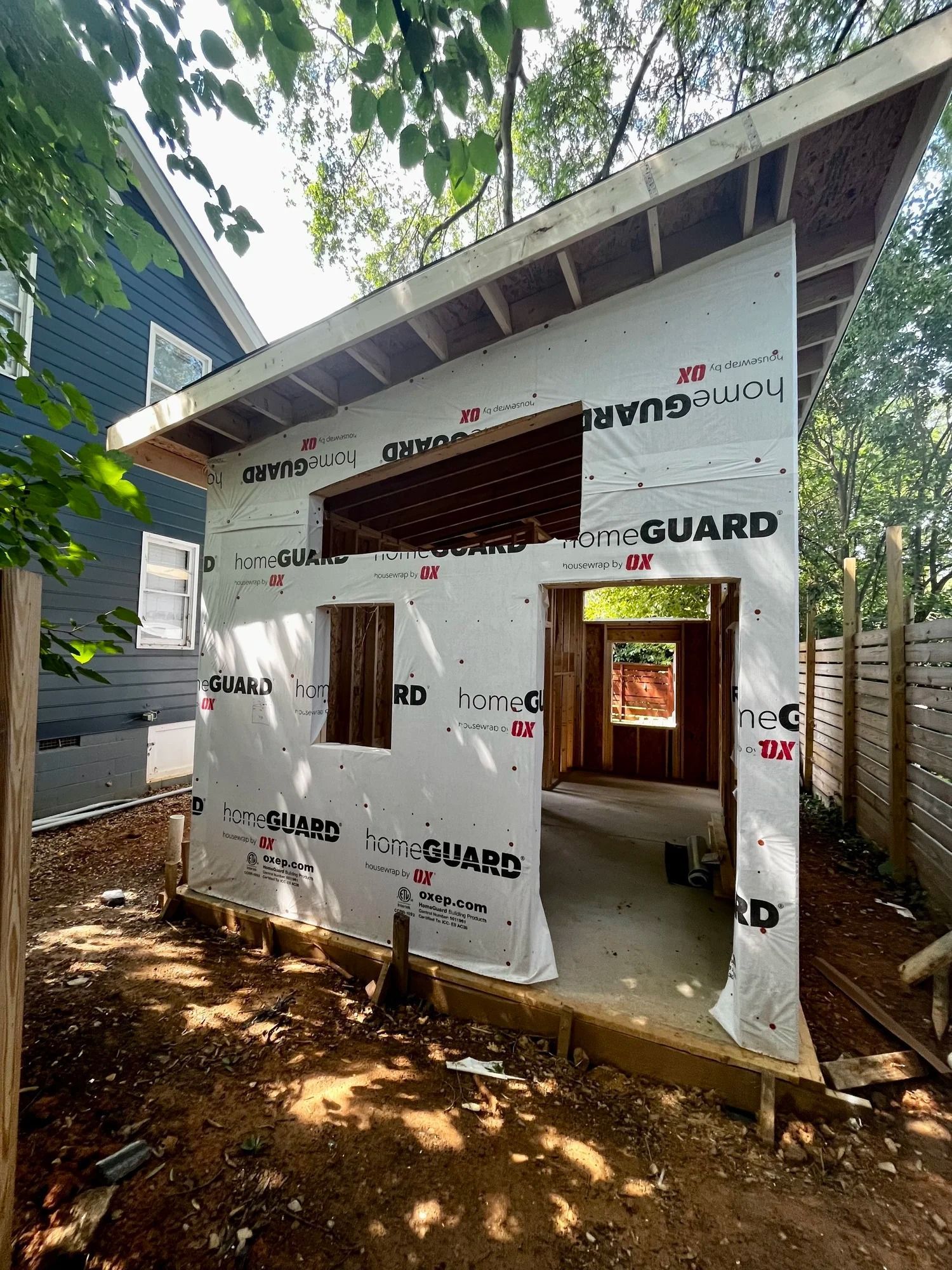 Small unfinished building wrapped in white construction sheeting under trees, with an open doorway and window.