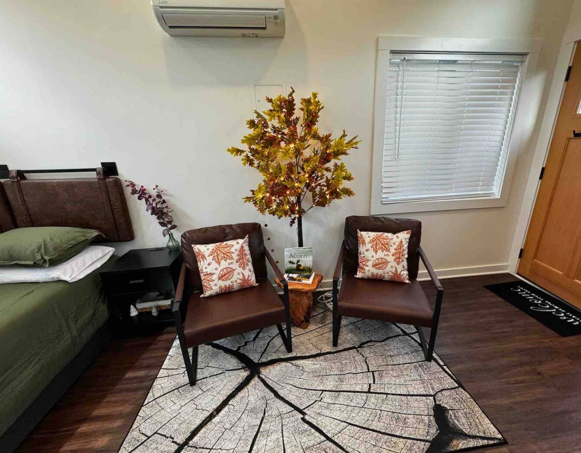 Cozy bedroom seating area with two brown chairs, leaf decor, rug, bed, and window blinds.