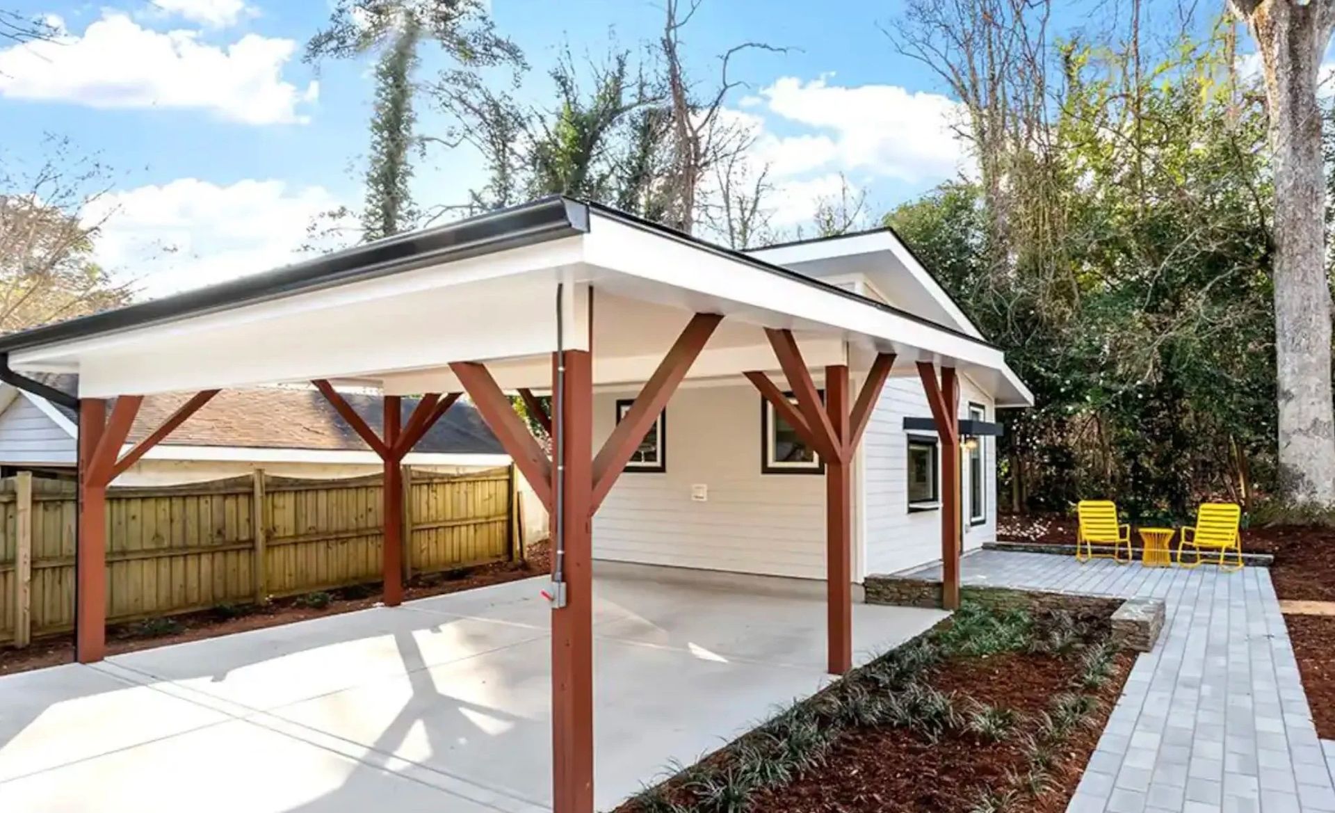 Covered backyard patio with white roof, brown posts, paved path, and yellow chairs beside a fence and trees