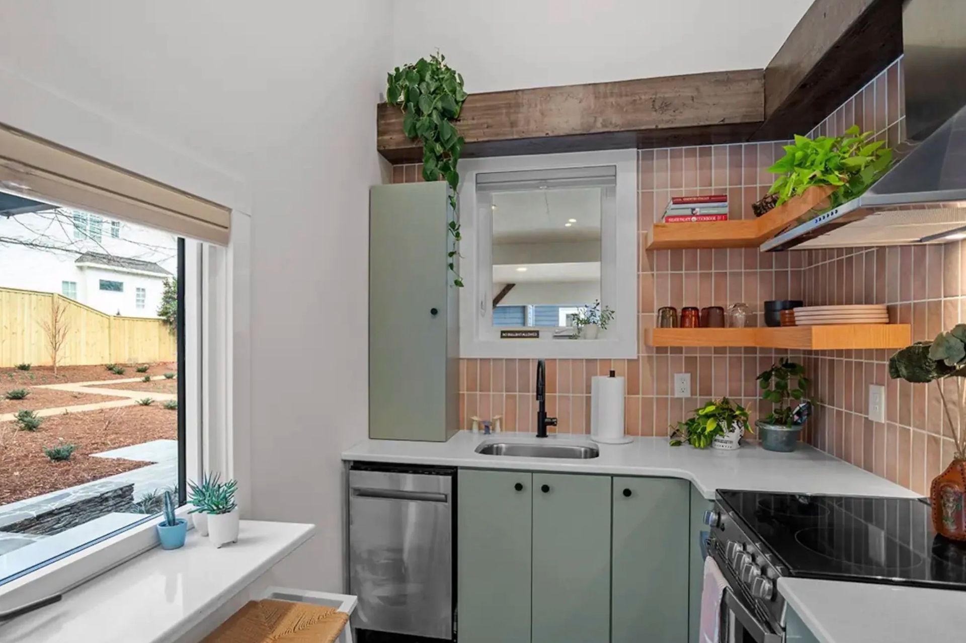 Small kitchen with sage cabinets, open wood shelves, mirror backsplash, and potted plants by a window