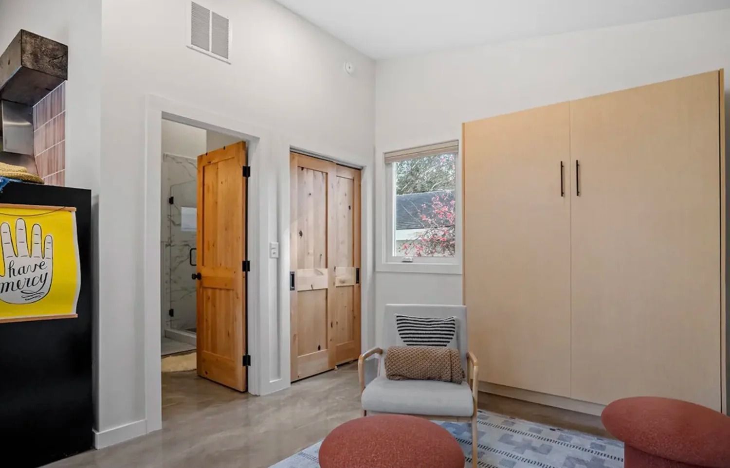 Bright bedroom with light wood wardrobe, open doors, window, and red ottoman chairs